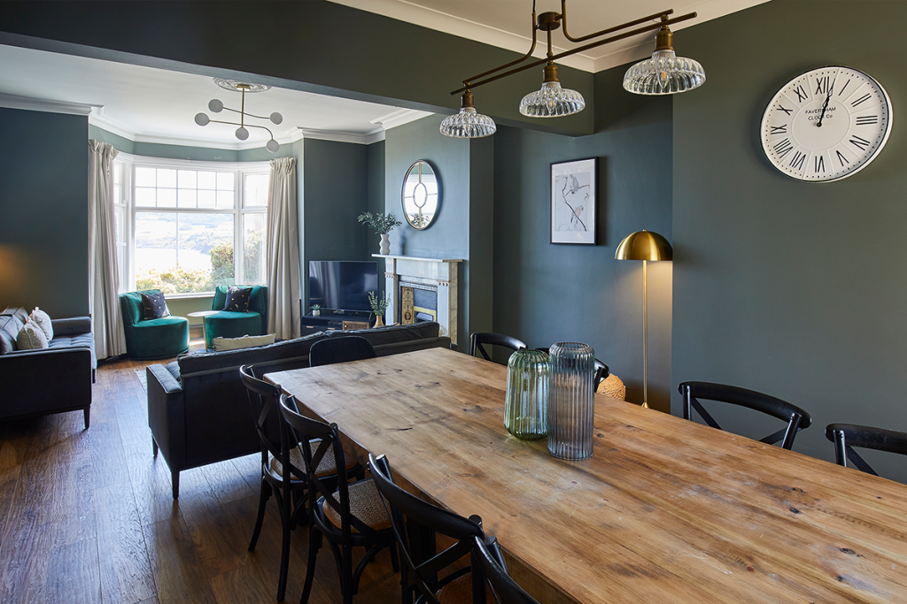 an oak dining table in a room with dark navy/black walls, brass features, and green chairs and sofa